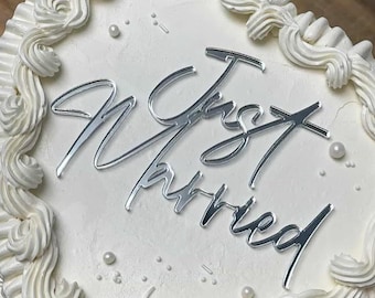 May include: A heart-shaped wedding cake with white frosting and decorative white frosting swirls. The cake is adorned with the words "Just Married" in silver script, and small white pearl sprinkles.
