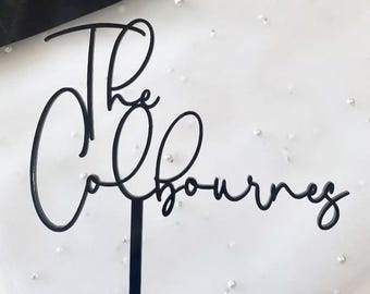 May include: Black cake topper with the words "The Colbournes" in elegant cursive script. The topper is mounted on a thin black stem, perfect for wedding or celebration cakes. The background is a white fabric with scattered pearls.