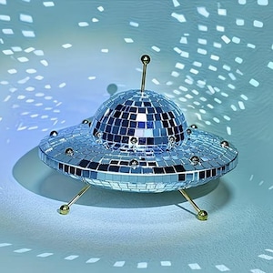 May include: A UFO-shaped decorative object covered in small, reflective silver mirror tiles. The structure features a central dome, a flat circular base, and three thin gold-toned metal legs. A single gold-toned antenna extends from the top centre.