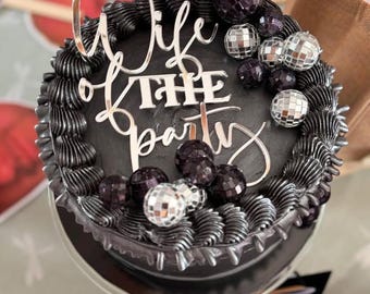 May include: A round cake with black frosting, featuring a silver cake topper that reads "Wife of THE party" in a mix of script and block lettering. The cake is decorated with small, reflective silver and dark purple disco ball ornaments.