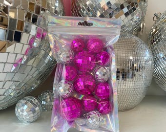 May include: A clear, iridescent plastic pouch containing a collection of small, round disco balls. The assortment includes several bright magenta pink mirrored spheres and a few silver mirrored spheres, all featuring a reflective tiled surface.