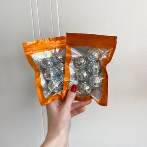 Pode incluir: Dois sacos laranja sel&aacute;veis com janelas transparentes, cada um contendo v&aacute;rias pequenas bolas de discoteca prateadas. Os sacos apresentam um forro interior hologr&aacute;fico que reflete a luz, exibindo as superf&iacute;cies espelhadas das bolas esf&eacute;ricas no interior.