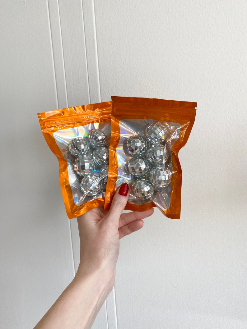 Pode incluir: Dois sacos laranja sel&aacute;veis com janelas transparentes, cada um contendo v&aacute;rias pequenas bolas de discoteca prateadas. Os sacos apresentam um forro interior hologr&aacute;fico que reflete a luz, exibindo as superf&iacute;cies espelhadas das bolas esf&eacute;ricas no interior.