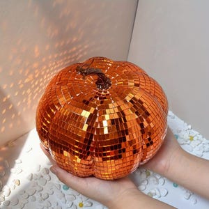 May include: A pumpkin-shaped decorative object covered in small, reflective orange mirror tiles. The surface is composed of a grid of square, metallic orange facets that catch and reflect light, creating a disco ball effect with a dark brown stem.