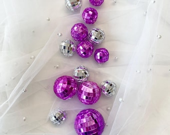 May include: A collection of spherical disco balls in varying sizes, featuring reflective silver mirrored tiles and vibrant purple mirrored tiles, arranged on a sheer white fabric surface decorated with small, scattered white pearls.