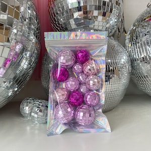 Pode incluir: Um saco de pl&aacute;stico transparente e iridescente com fecho, contendo uma cole&ccedil;&atilde;o de pequenas bolas de discoteca esf&eacute;ricas em v&aacute;rios tons de rosa e magenta. Cada bola &eacute; coberta por min&uacute;sculos azulejos de espelho quadrados e refletores.