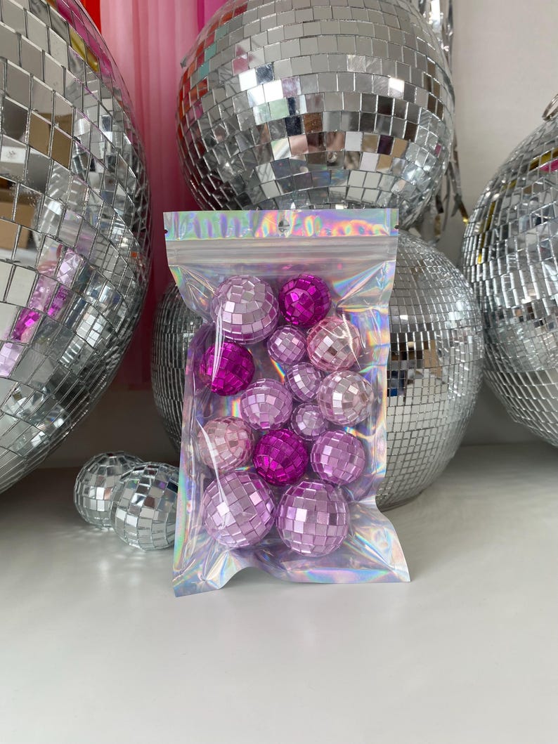 Pode incluir: Um saco de pl&aacute;stico transparente e iridescente com fecho, contendo uma cole&ccedil;&atilde;o de pequenas bolas de discoteca esf&eacute;ricas em v&aacute;rios tons de rosa e magenta. Cada bola &eacute; coberta por min&uacute;sculos azulejos de espelho quadrados e refletores.