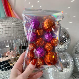 Pode incluir: Um saco de pl&aacute;stico iridescente transparente contendo uma cole&ccedil;&atilde;o de pequenas bolas de discoteca esf&eacute;ricas em tons vibrantes de laranja e roxo. Cada bola &eacute; coberta por pequenos espelhos quadrados refletores que captam a luz.