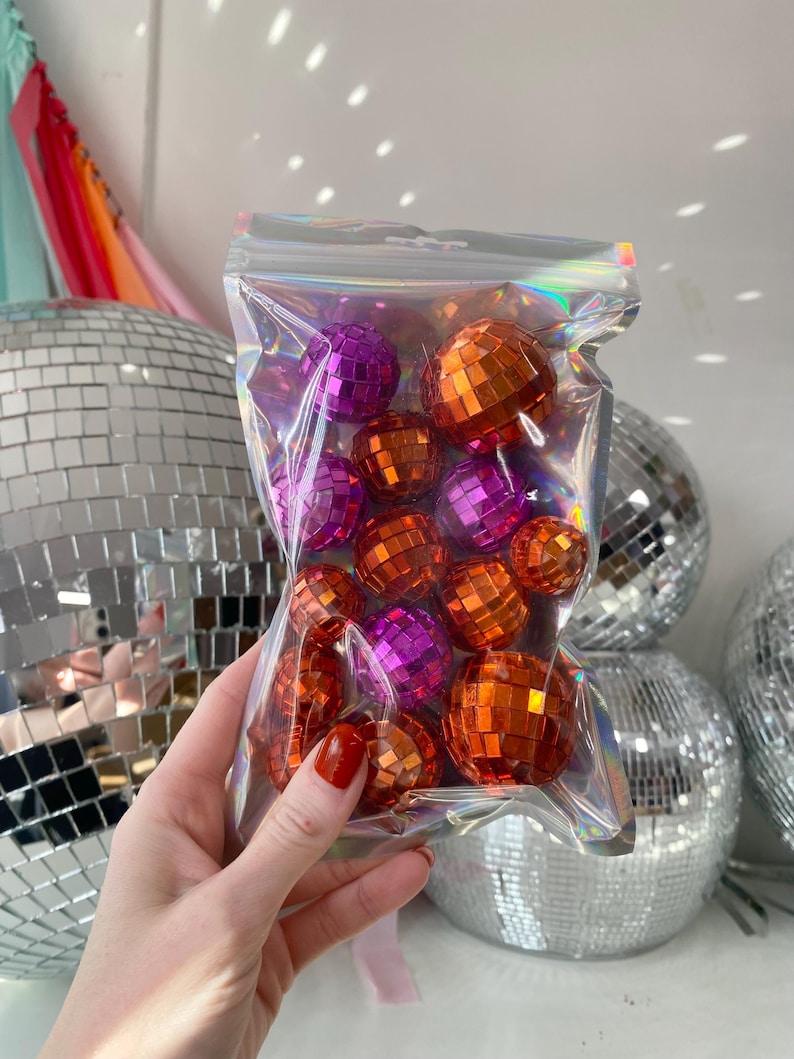 Pode incluir: Um saco de pl&aacute;stico iridescente transparente contendo uma cole&ccedil;&atilde;o de pequenas bolas de discoteca esf&eacute;ricas em tons vibrantes de laranja e roxo. Cada bola &eacute; coberta por pequenos espelhos quadrados refletores que captam a luz.