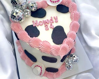 May include: A heart-shaped cake with white frosting, black cow-print spots, and pink piped borders. The top features the text "Howdy 24" in pink, along with miniature silver disco balls and a small white cowboy hat with pink accents.
