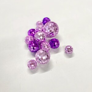 Puede incluir: Una colecci&oacute;n de peque&ntilde;as bolas de discoteca esf&eacute;ricas en varios tonos de morado y lavanda claro. Cada bola est&aacute; cubierta con diminutos azulejos de espejo cuadrados y reflectantes que crean una superficie facetada de diferentes tama&ntilde;os.