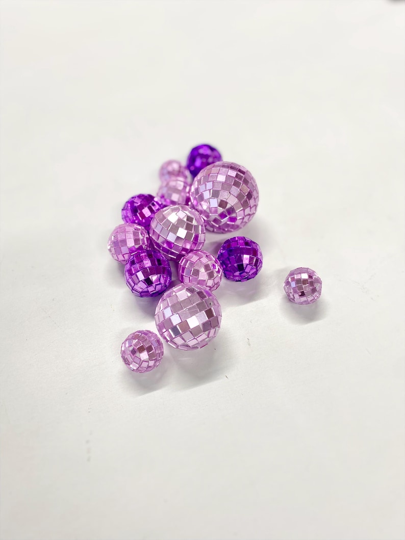 Puede incluir: Una colecci&oacute;n de peque&ntilde;as bolas de discoteca esf&eacute;ricas en varios tonos de morado y lavanda claro. Cada bola est&aacute; cubierta con diminutos azulejos de espejo cuadrados y reflectantes que crean una superficie facetada de diferentes tama&ntilde;os.