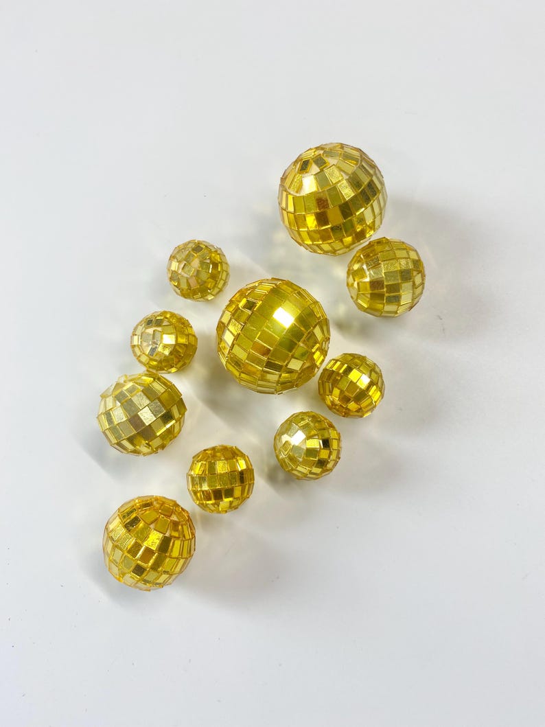 Puede incluir: Una colecci&oacute;n de diez bolas de discoteca esf&eacute;ricas de varios tama&ntilde;os, cubiertas con peque&ntilde;os azulejos de espejo cuadrados de color dorado reflectante. Las esferas est&aacute;n dispuestas en un patr&oacute;n disperso con un acabado met&aacute;lico.