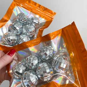 Pode incluir: Dois sacos de pl&aacute;stico laranja iridescente com fecho, cada um contendo v&aacute;rias pequenas bolas de discoteca esf&eacute;ricas cobertas com min&uacute;sculos espelhos prateados refletores. Os sacos s&atilde;o transparentes no centro, exibindo as superf&iacute;cies brilhantes e facetadas.