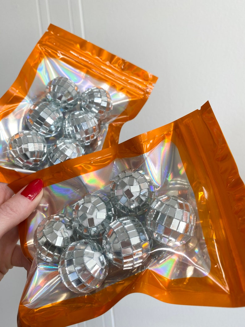 Pode incluir: Dois sacos de pl&aacute;stico laranja iridescente com fecho, cada um contendo v&aacute;rias pequenas bolas de discoteca esf&eacute;ricas cobertas com min&uacute;sculos espelhos prateados refletores. Os sacos s&atilde;o transparentes no centro, exibindo as superf&iacute;cies brilhantes e facetadas.