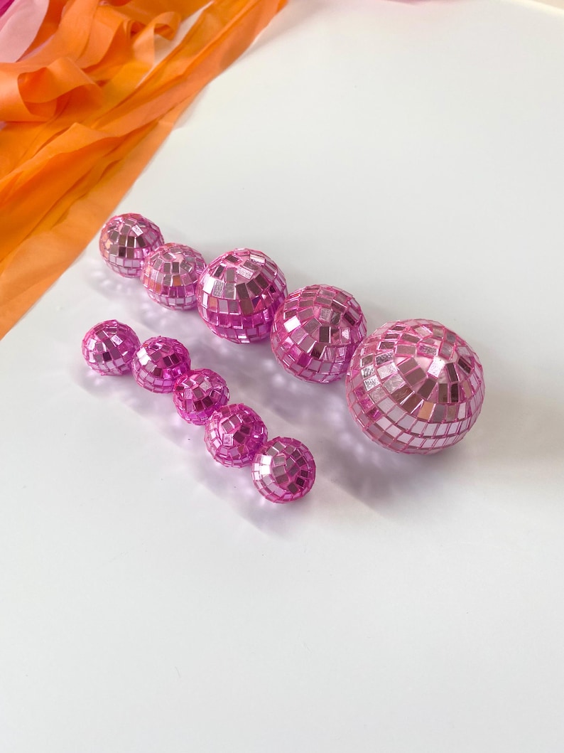 Puede incluir: Un conjunto de nueve bolas de discoteca esf&eacute;ricas de distintos tama&ntilde;os, cubiertas con peque&ntilde;os espejos cuadrados de color rosa reflectante. Las esferas est&aacute;n dispuestas en dos filas y presentan un acabado rosa met&aacute;lico brillante.