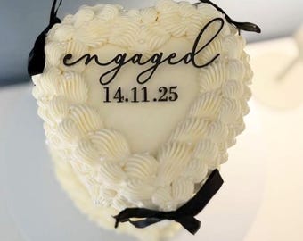 May include: A heart-shaped cake with white frosting and decorative swirls. The cake has the word "engaged" and the date "14.11.25" written on it. Black ribbons are tied around the cake.