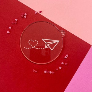 Può includere: Un cerchio acrilico trasparente con un aeroplano di carta bianco e una linea punteggiata che porta a un cuore. Il cerchio è su una superficie rossa con cuori rosa sparsi intorno.