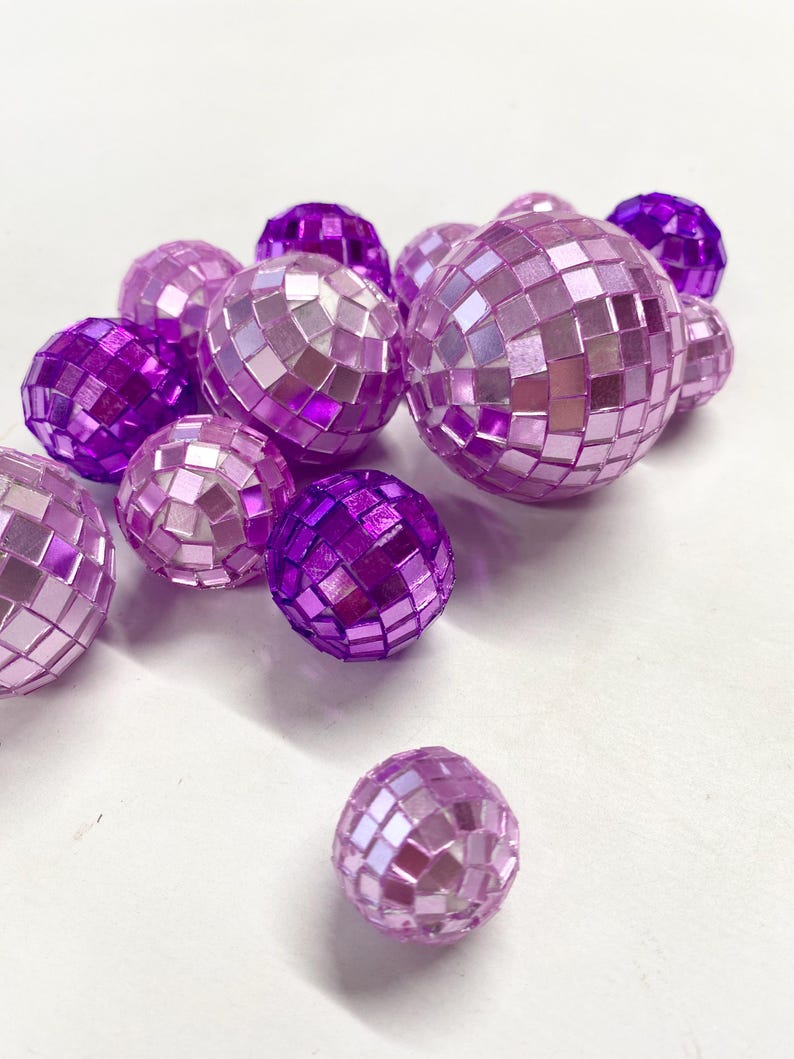 Puede incluir: Una colecci&oacute;n de bolas de discoteca esf&eacute;ricas en varios tonos de morado y rosa claro. Cada bola est&aacute; cubierta con peque&ntilde;os azulejos de espejo cuadrados y reflectantes que crean una superficie facetada. Las esferas var&iacute;an en tama&ntilde;o y grupo.