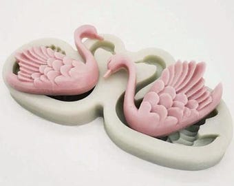 May include: A light gray silicone mold featuring two recessed swan shapes. Each swan cavity displays detailed feather textures on the wings and a curved neck. The mold is designed to create two identical swan-shaped figures in a side-by-side orientation.