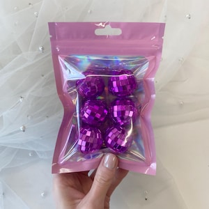 Pode incluir: Um conjunto de seis pequenas bolas de discoteca roxas com uma superf&iacute;cie espelhada refletora, embaladas numa bolsa de pl&aacute;stico transparente e iridescente com uma borda rosa e um orif&iacute;cio superior para pendurar.
