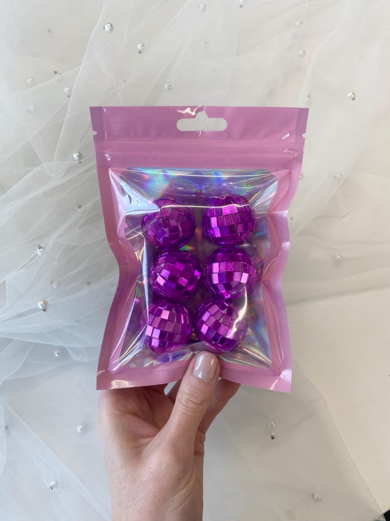 Pode incluir: Um conjunto de seis pequenas bolas de discoteca roxas com uma superf&iacute;cie espelhada refletora, embaladas numa bolsa de pl&aacute;stico transparente e iridescente com uma borda rosa e um orif&iacute;cio superior para pendurar.