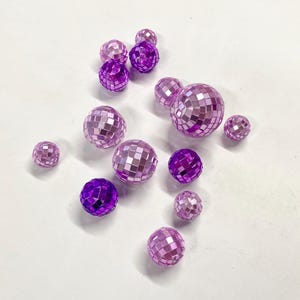 Puede incluir: Una colecci&oacute;n de doce bolas de discoteca esf&eacute;ricas de varios tama&ntilde;os, con una superficie reflectante compuesta por peque&ntilde;os azulejos de espejo cuadrados de color morado y morado claro, dispuestas de forma dispersa sobre una superficie blanca.