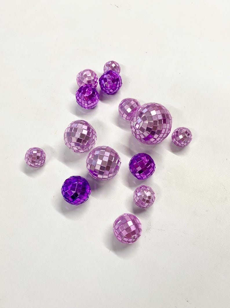 Puede incluir: Una colecci&oacute;n de doce bolas de discoteca esf&eacute;ricas de varios tama&ntilde;os, con una superficie reflectante compuesta por peque&ntilde;os azulejos de espejo cuadrados de color morado y morado claro, dispuestas de forma dispersa sobre una superficie blanca.