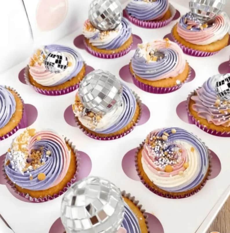 Pode incluir: Uma cole&ccedil;&atilde;o de cupcakes dispostos numa caixa branca, cada um com um redemoinho de cobertura em tons pastel de roxo, rosa e branco. Alguns cupcakes s&atilde;o decorados com pequenos ornamentos de bola de discoteca prateados e outros com granulados dourados.