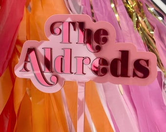 May include: A custom cake topper featuring the text The Aldreds in a dark pink reflective script font. The lettering is mounted on a light pink, cloud-shaped acrylic backing with a single vertical stem extending from the bottom center.