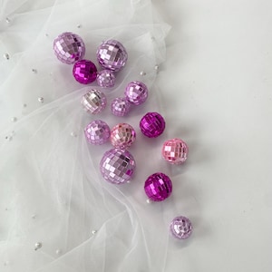 Pode incluir: Uma cole&ccedil;&atilde;o de bolas de discoteca esf&eacute;ricas em v&aacute;rios tons de rosa e roxo, variando do lavanda claro ao magenta profundo. Cada bola &eacute; coberta com pequenos espelhos quadrados reflexivos que captam a luz, criando uma superf&iacute;cie facetada.