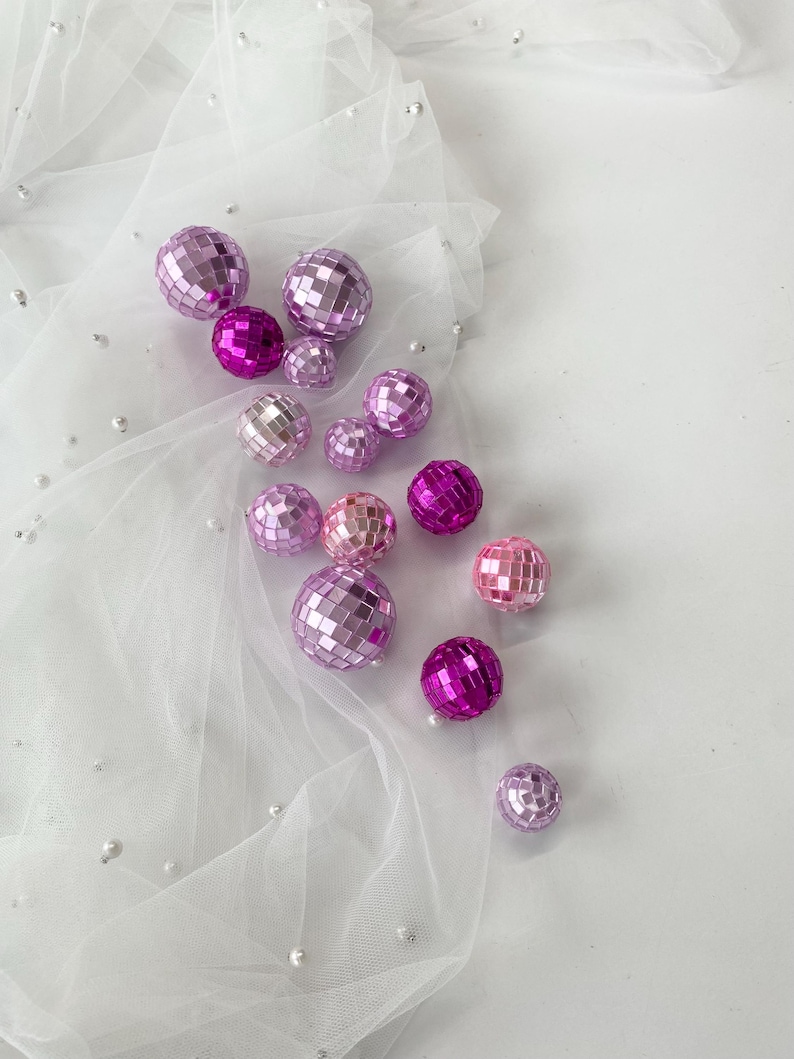 Pode incluir: Uma cole&ccedil;&atilde;o de bolas de discoteca esf&eacute;ricas em v&aacute;rios tons de rosa e roxo, variando do lavanda claro ao magenta profundo. Cada bola &eacute; coberta com pequenos espelhos quadrados reflexivos que captam a luz, criando uma superf&iacute;cie facetada.