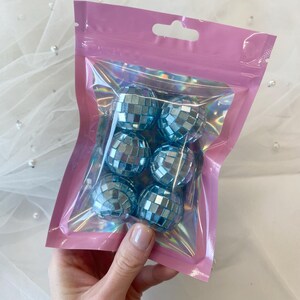 Pode incluir: Um conjunto de seis pequenas bolas de discoteca esf&eacute;ricas com um acabamento refletor azul-claro, cada uma coberta por pequenos espelhos quadrados. As bolas est&atilde;o contidas dentro de uma bolsa de pl&aacute;stico transparente e iridescente.