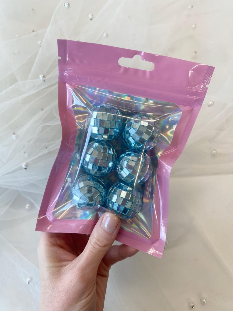 Pode incluir: Um conjunto de seis pequenas bolas de discoteca esf&eacute;ricas com um acabamento refletor azul-claro, cada uma coberta por pequenos espelhos quadrados. As bolas est&atilde;o contidas dentro de uma bolsa de pl&aacute;stico transparente e iridescente.