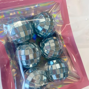 Pode incluir: Um conjunto de cinco ornamentos esf&eacute;ricos de bola de discoteca com azulejos quadrados espelhados em azul claro. Os ornamentos est&atilde;o contidos numa bolsa de pl&aacute;stico transparente e iridescente com uma borda rosa e um orif&iacute;cio para pendurar.