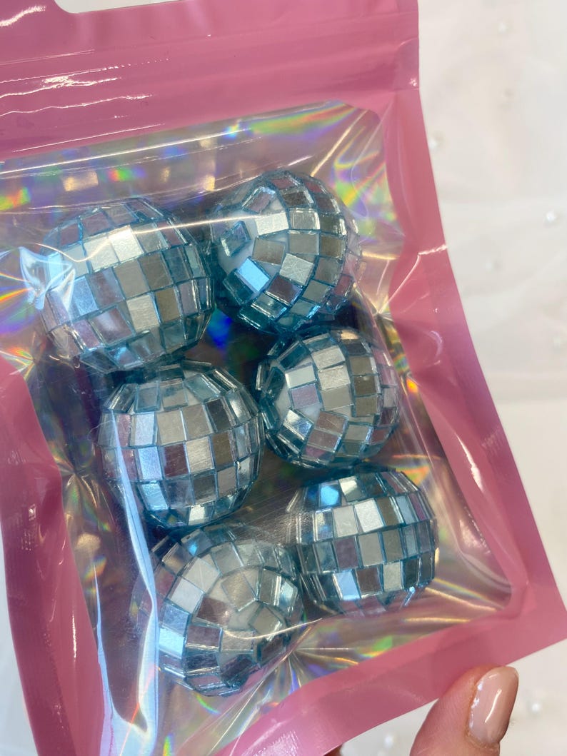 Pode incluir: Um conjunto de cinco ornamentos esf&eacute;ricos de bola de discoteca com azulejos quadrados espelhados em azul claro. Os ornamentos est&atilde;o contidos numa bolsa de pl&aacute;stico transparente e iridescente com uma borda rosa e um orif&iacute;cio para pendurar.