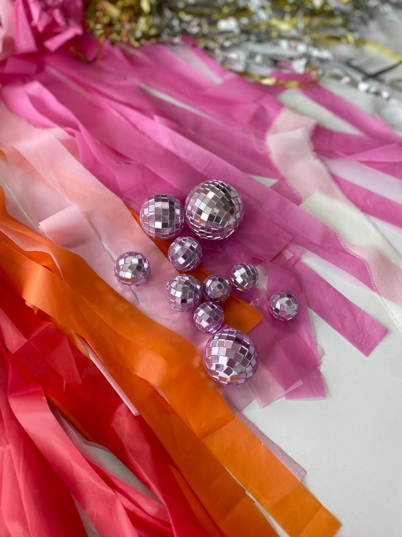 Puede incluir: Una colecci&oacute;n de peque&ntilde;as bolas de discoteca esf&eacute;ricas de color morado claro con facetas de espejo reflectantes, dispuestas sobre un fondo de largas tiras de serpentinas de papel rosa, naranja y blanco, con algunos acentos met&aacute;licos dorados y plateados.