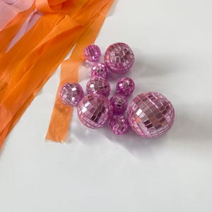 Puede incluir: Una colecci&oacute;n de diez bolas de discoteca rosas de varios tama&ntilde;os, cada una cubierta con peque&ntilde;os espejos cuadrados reflectantes. Las esferas est&aacute;n dispuestas en un grupo junto a varias tiras largas y fluidas de cinta de color naranja y rosa.