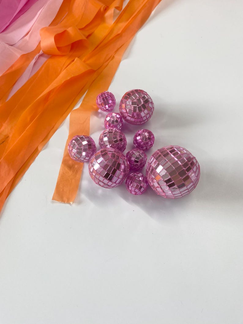 Puede incluir: Una colecci&oacute;n de diez bolas de discoteca rosas de varios tama&ntilde;os, cada una cubierta con peque&ntilde;os espejos cuadrados reflectantes. Las esferas est&aacute;n dispuestas en un grupo junto a varias tiras largas y fluidas de cinta de color naranja y rosa.