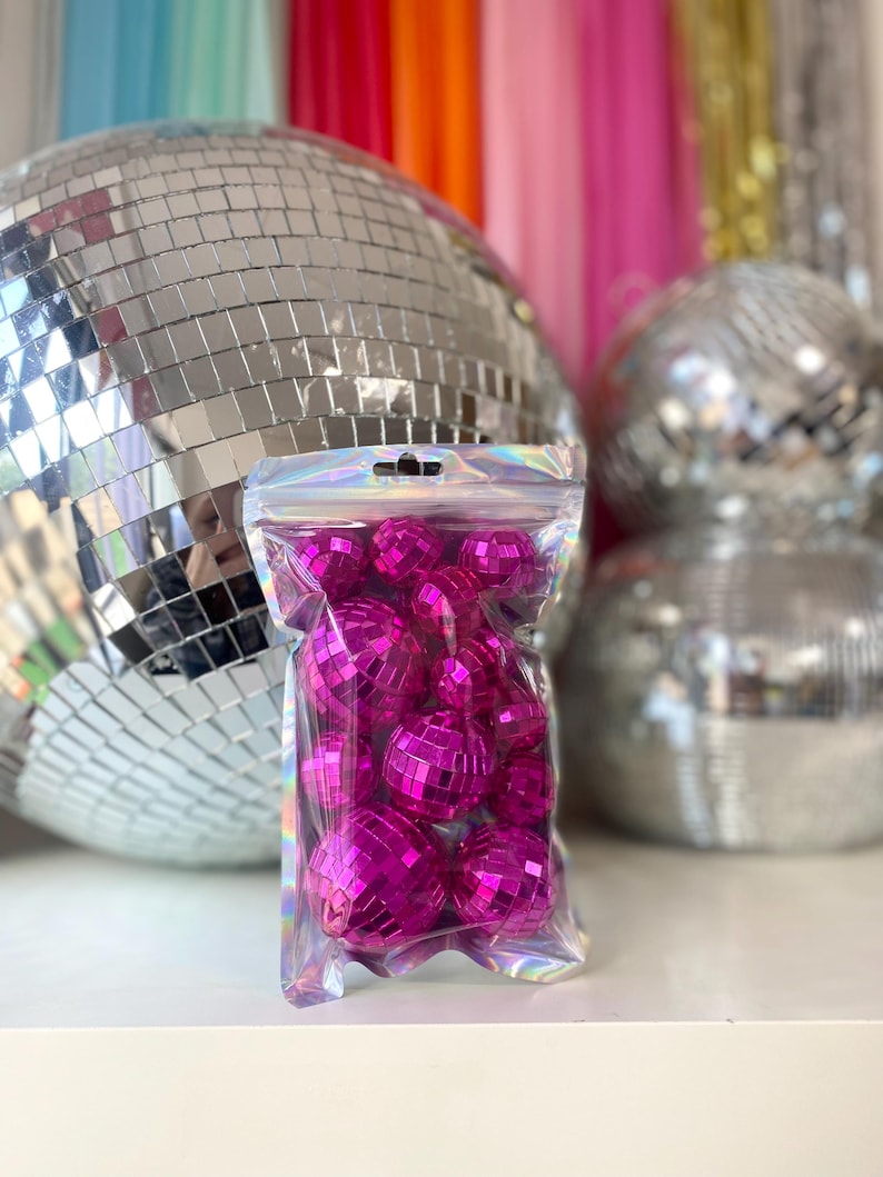 Pode incluir: Um saco de pl&aacute;stico transparente e iridescente com fecho, contendo uma cole&ccedil;&atilde;o de pequenas bolas de discoteca redondas em cor-de-rosa choque. Cada bola est&aacute; coberta por min&uacute;sculos espelhos quadrados refletores que captam a luz.