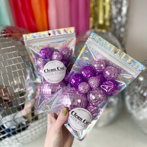 Puede incluir: Dos bolsas hologr&aacute;ficas transparentes resellables que contienen m&uacute;ltiples adornos de bolas de discoteca moradas con facetas de espejo. Cada bolsa tiene una etiqueta circular blanca con el texto Clean Cut LASER STUDIO y se sujeta el asa.