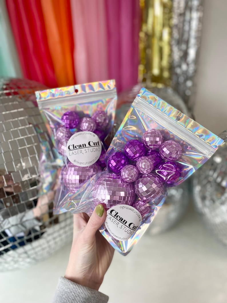 Puede incluir: Dos bolsas hologr&aacute;ficas transparentes resellables que contienen m&uacute;ltiples adornos de bolas de discoteca moradas con facetas de espejo. Cada bolsa tiene una etiqueta circular blanca con el texto Clean Cut LASER STUDIO y se sujeta el asa.