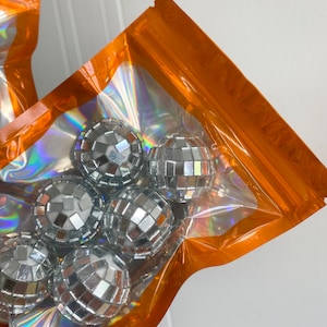 Pode incluir: Um conjunto de pequenas bolas de discoteca esf&eacute;ricas cobertas com espelhos prateados refletivos, embaladas dentro de uma bolsa de pl&aacute;stico transparente com uma borda laranja vibrante e um acabamento de filme hologr&aacute;fico iridescente.