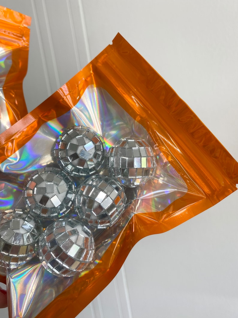 Pode incluir: Um conjunto de pequenas bolas de discoteca esf&eacute;ricas cobertas com espelhos prateados refletivos, embaladas dentro de uma bolsa de pl&aacute;stico transparente com uma borda laranja vibrante e um acabamento de filme hologr&aacute;fico iridescente.