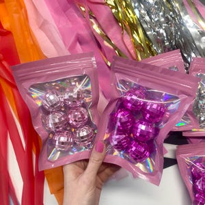 Pode incluir: Dois sacos cor-de-rosa sel&aacute;veis contendo pequenas bolas de discoteca refletoras. Um saco cont&eacute;m esferas espelhadas cor-de-rosa claro, enquanto o outro cont&eacute;m esferas espelhadas cor-de-rosa choque. Os sacos t&ecirc;m uma janela transparente e detalhes hologr&aacute;ficos.