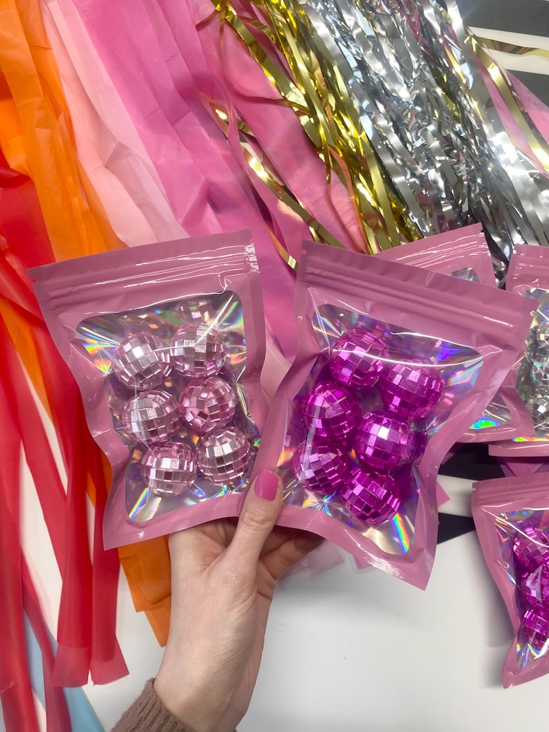 Pode incluir: Dois sacos cor-de-rosa sel&aacute;veis contendo pequenas bolas de discoteca refletoras. Um saco cont&eacute;m esferas espelhadas cor-de-rosa claro, enquanto o outro cont&eacute;m esferas espelhadas cor-de-rosa choque. Os sacos t&ecirc;m uma janela transparente e detalhes hologr&aacute;ficos.