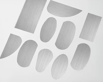 May include: A collection of nine stainless steel cake scrapers and icing smoothers in various shapes, including rectangular, oval, and semi-circular designs. Some pieces feature serrated edges for texturing, while others have smooth, straight edges.