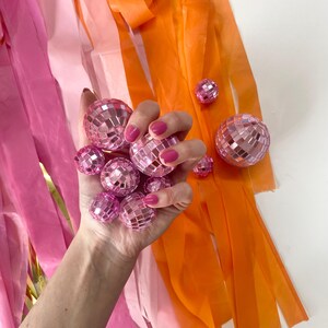 Puede incluir: Una colecci&oacute;n de peque&ntilde;as bolas de discoteca rosas con superficie facetada, sostenidas juntas en un grupo. Largas y finas tiras de serpentinas de pl&aacute;stico rosa, rosa claro y naranja cuelgan verticalmente detr&aacute;s de las bolas.