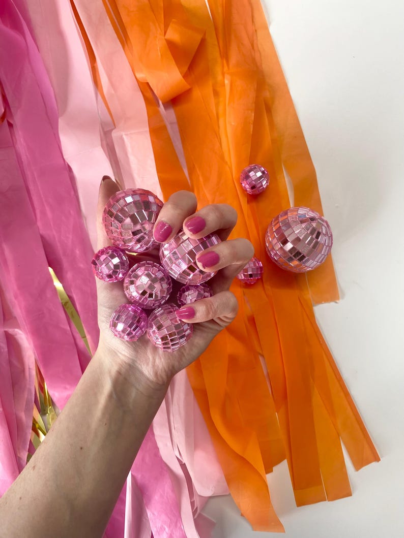 Puede incluir: Una colecci&oacute;n de peque&ntilde;as bolas de discoteca rosas con superficie facetada, sostenidas juntas en un grupo. Largas y finas tiras de serpentinas de pl&aacute;stico rosa, rosa claro y naranja cuelgan verticalmente detr&aacute;s de las bolas.