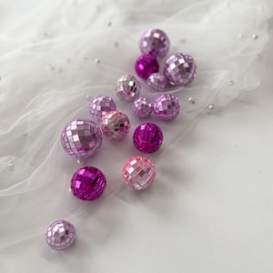 Pode incluir: Uma cole&ccedil;&atilde;o de pequenas bolas de discoteca esf&eacute;ricas em v&aacute;rios tons de rosa e roxo, variando do lavanda claro ao magenta profundo. Cada bola &eacute; coberta por min&uacute;sculos espelhos quadrados reflexivos que criam uma superf&iacute;cie facetada.